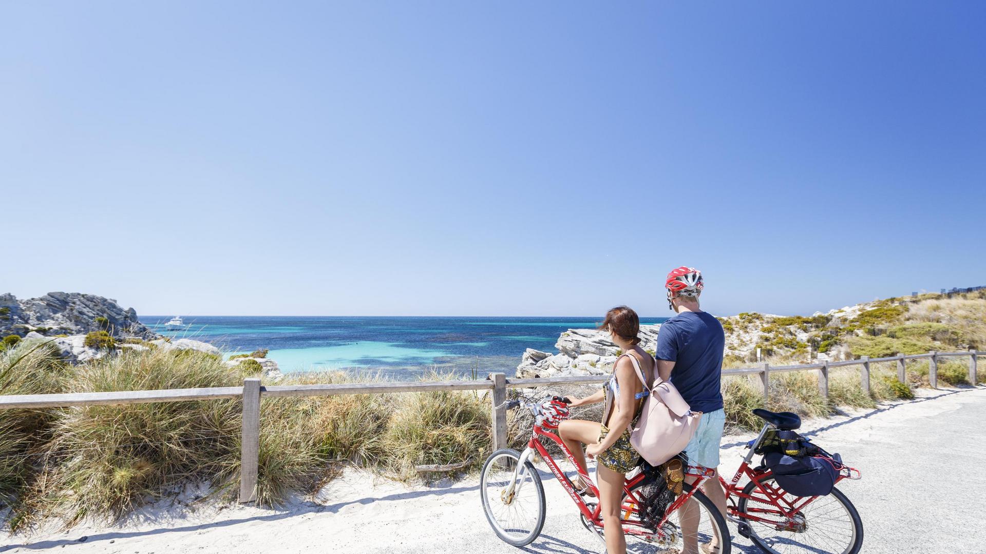 Two people stand beside a red bicycle on a coastal path overlooking a bay at Rottnest Island.