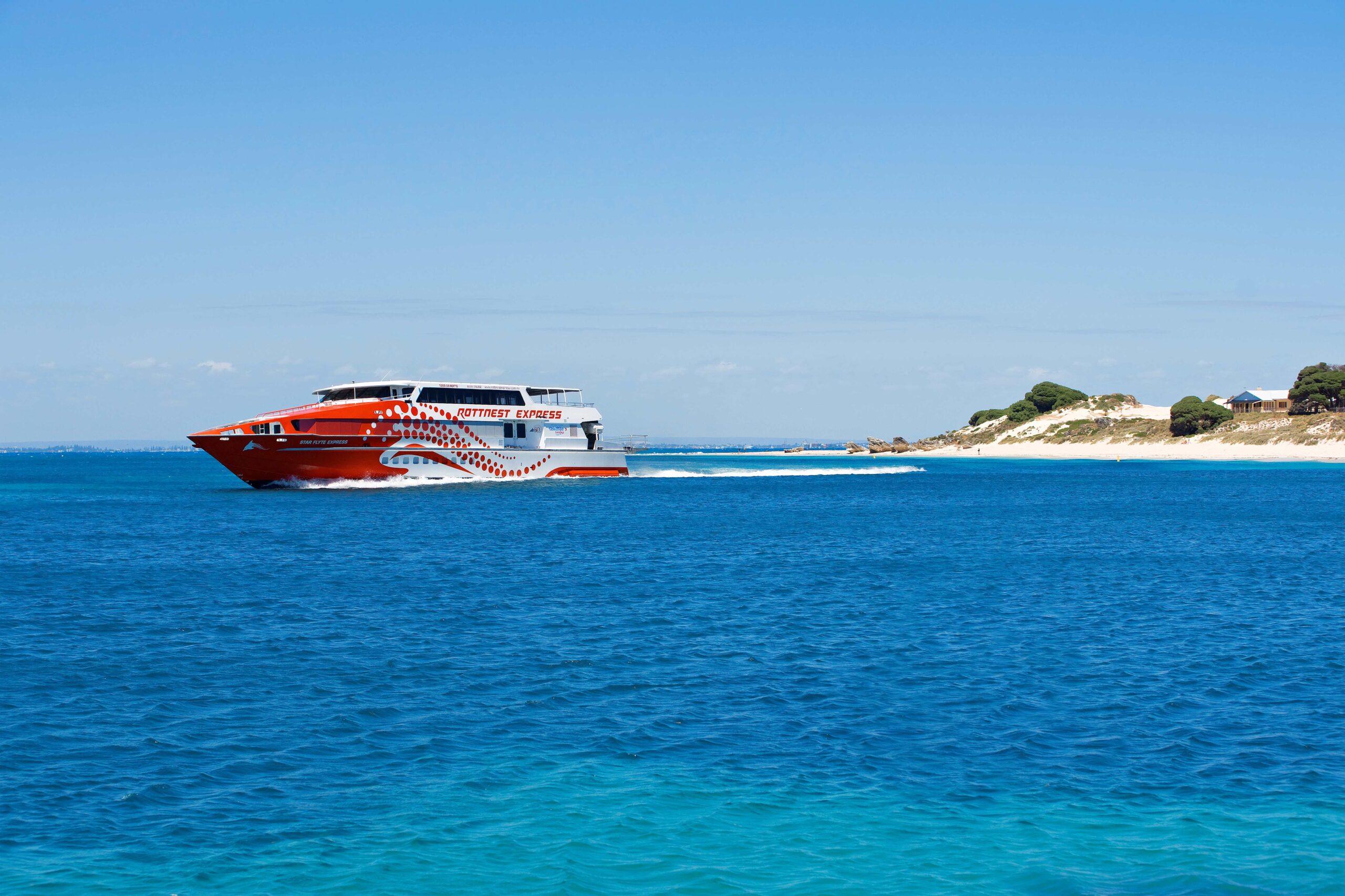 A red and white ferry cruises crystal clear waters and arrives at Rottnest Island under bright blue skies, there is a small sandbank visible in the background.