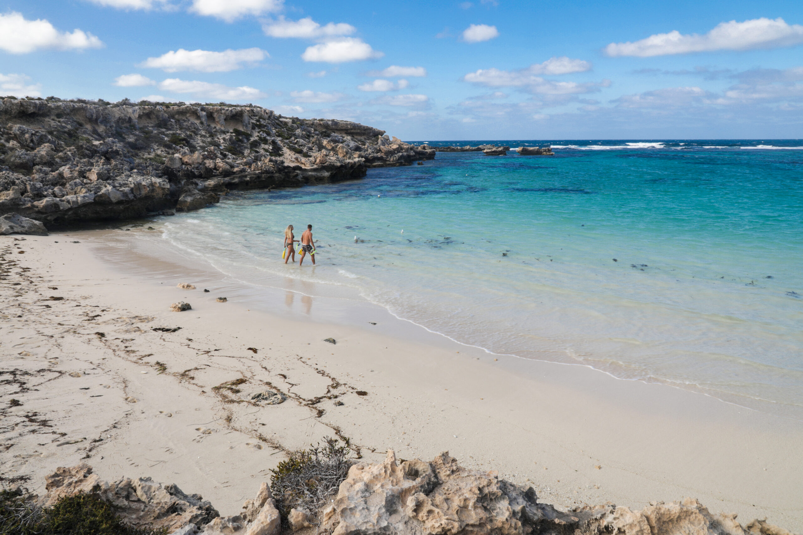 A few people swim and relax under a bright blue sky with soft white clouds at Little Salmon Bay at Rottnest Island.