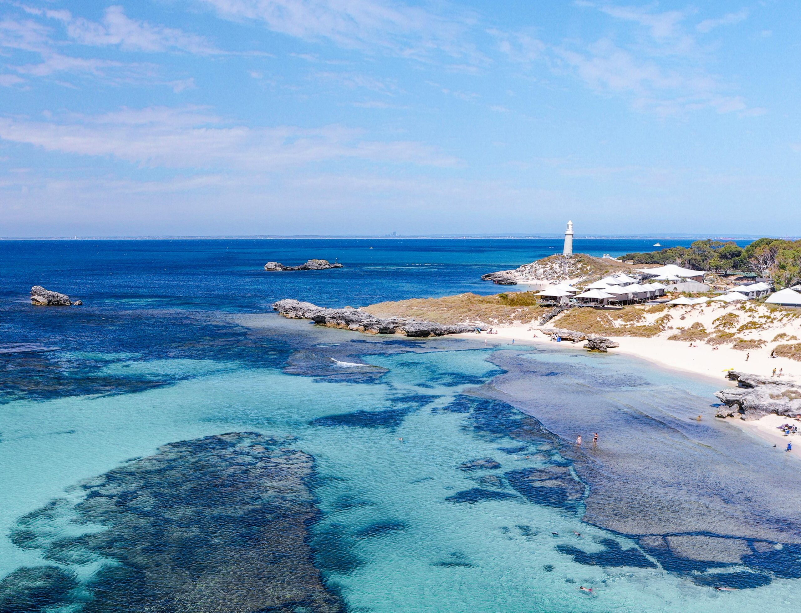 People swim and relax along The Basin at Rottnest Island, and a lighthouse is perched on a low dune under a clear blue sky.