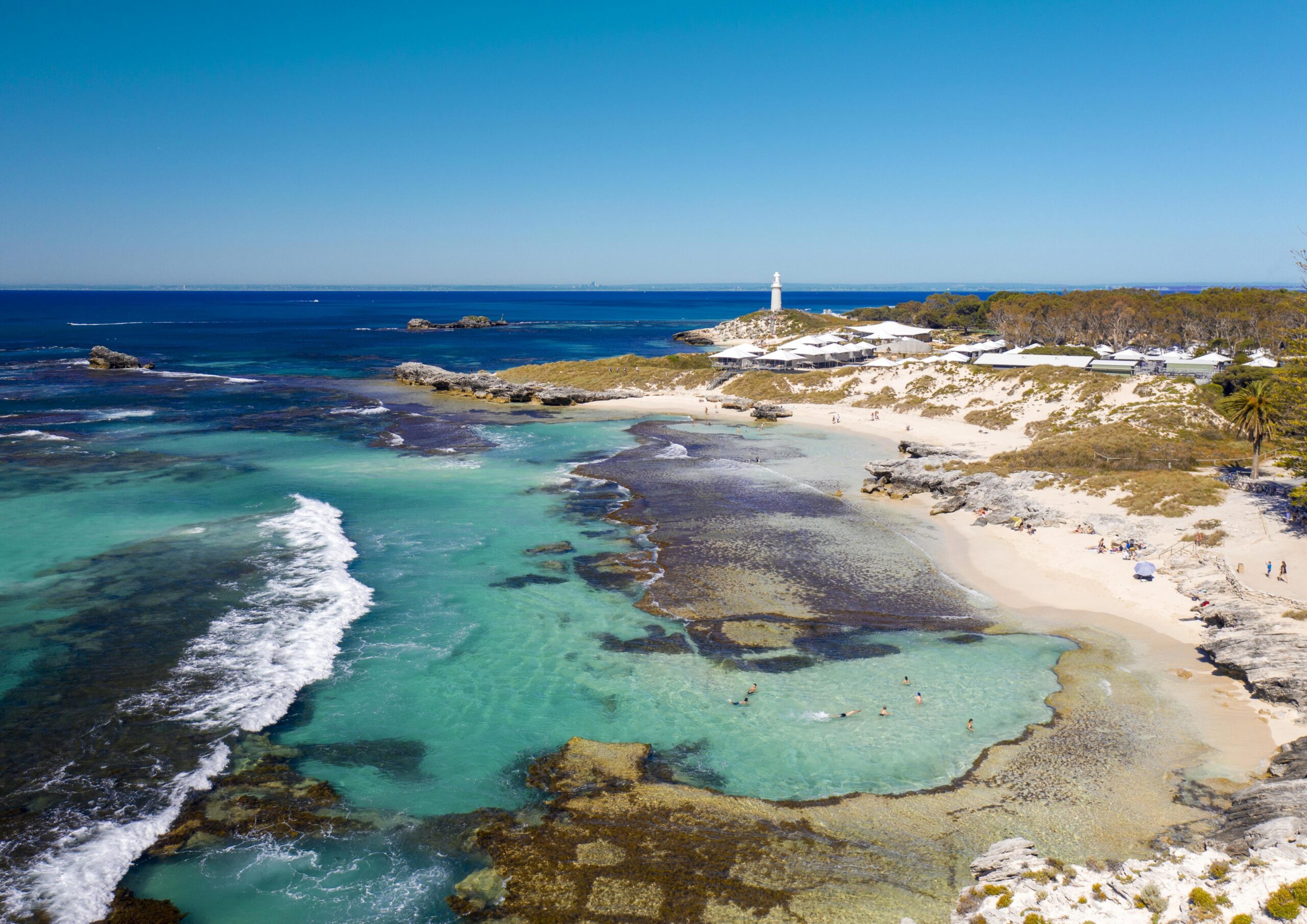 Turquoise clear water laps at a sandy beach with coral reefs and rock formations visible beneath the surface. A white lighthouse sits atop a rocky outcrop in the distance.