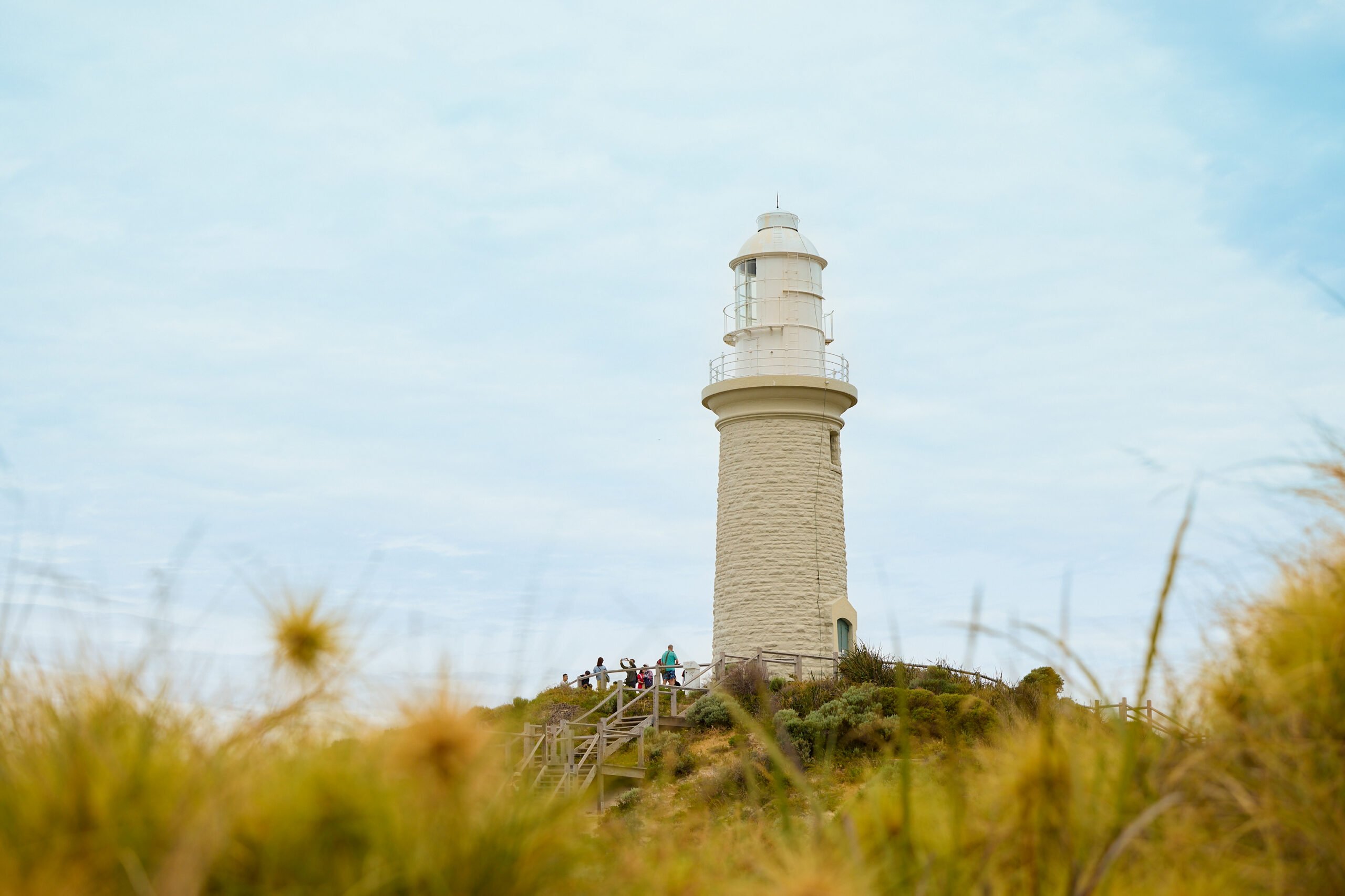 A lighthouse rises from a grassy hill, with a wooden walkway leading up to it and several people gathered at its base beneath a softly clouded sky.