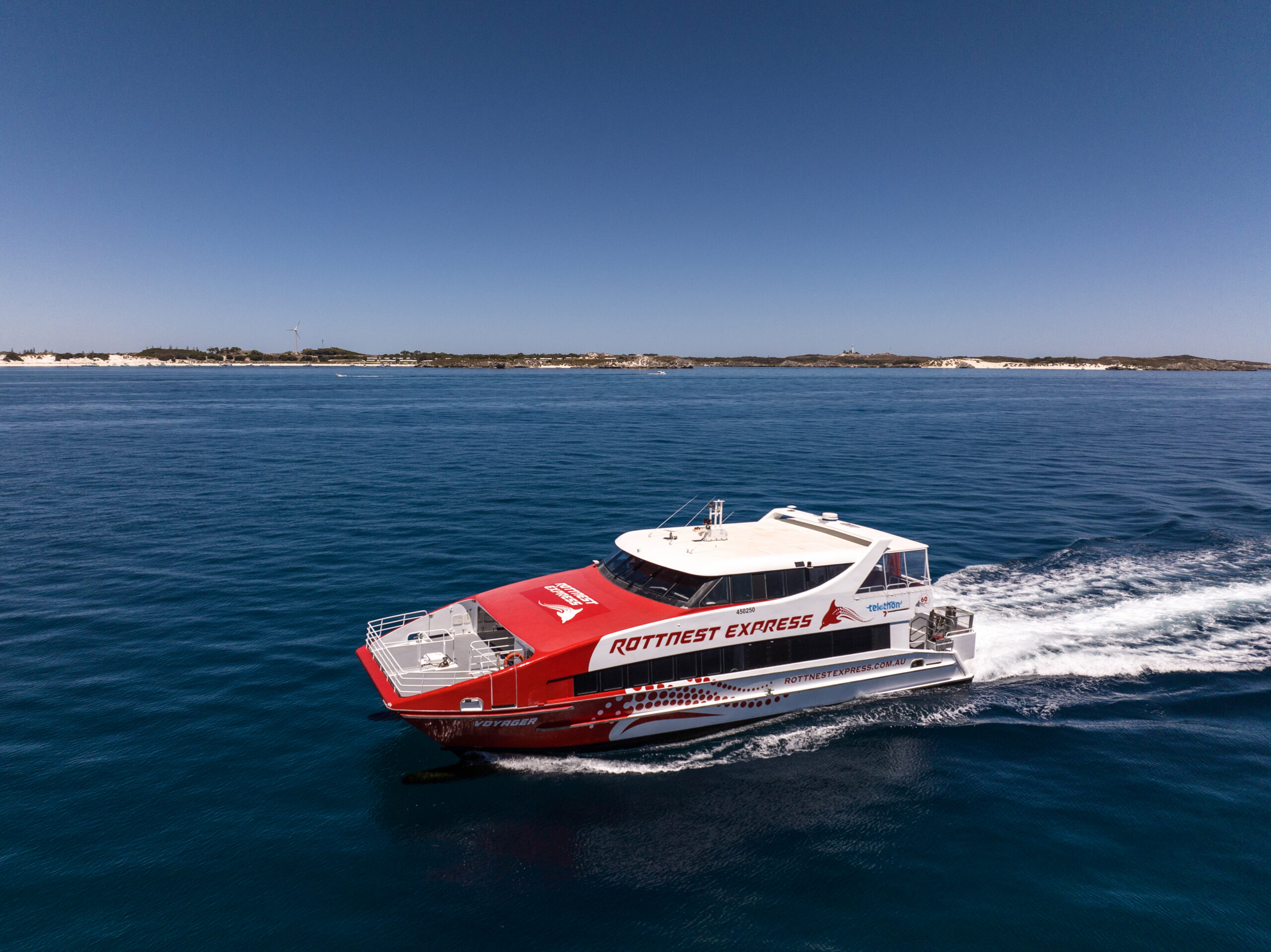 A red and white Rottnest Express ferry moves across calm blue water, with a sandy coastline and a single wind turbine visible beneath a clear sky.